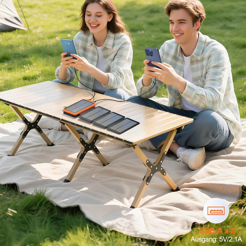 Solar-Powerbank auf einem Campingtisch lädt zwei Smartphones, daneben zwei Personen auf einer Decke